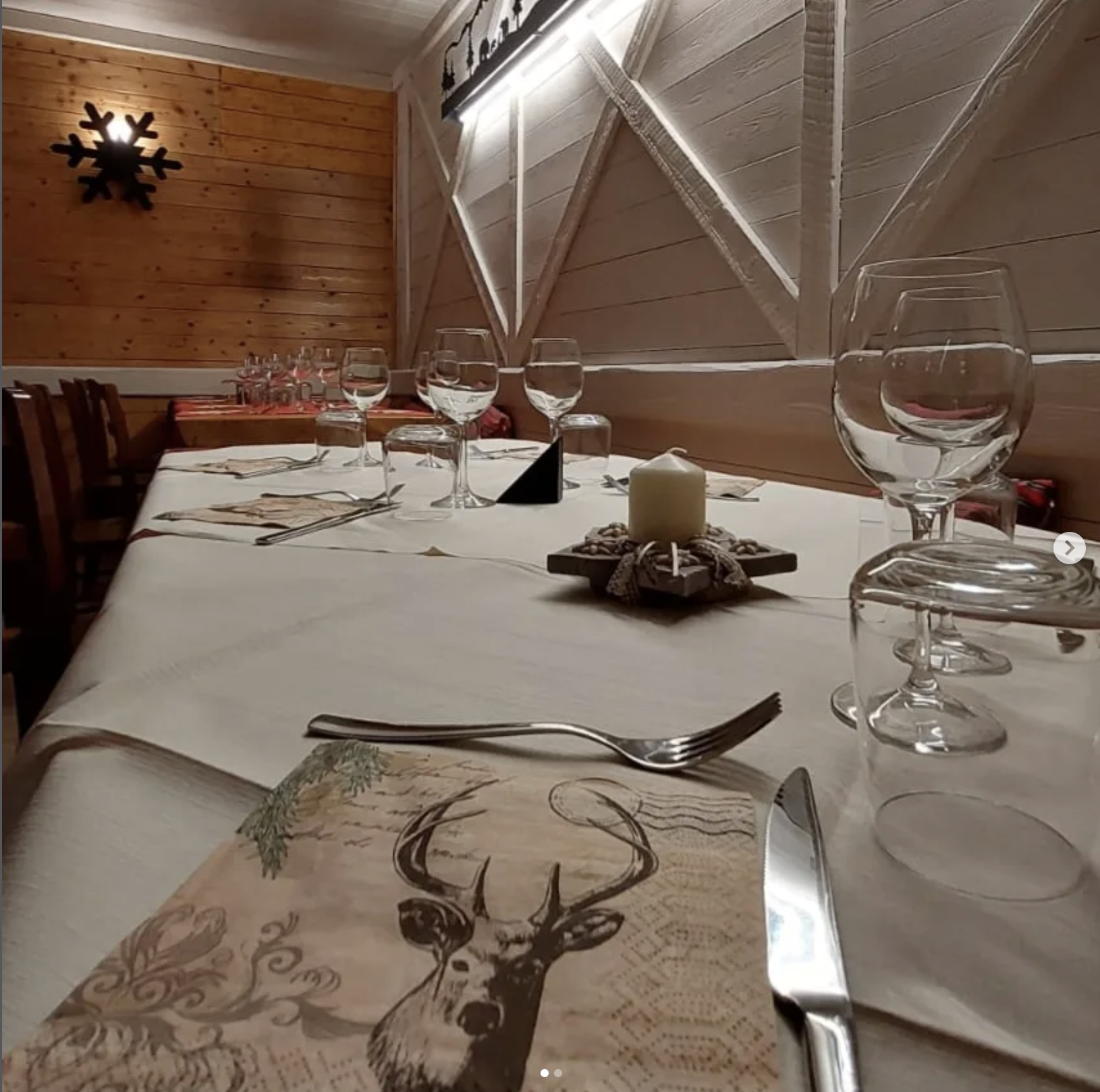La sala della baita apparecchiata: tavolo lungo, calici, candela e tovaglioli col cervo